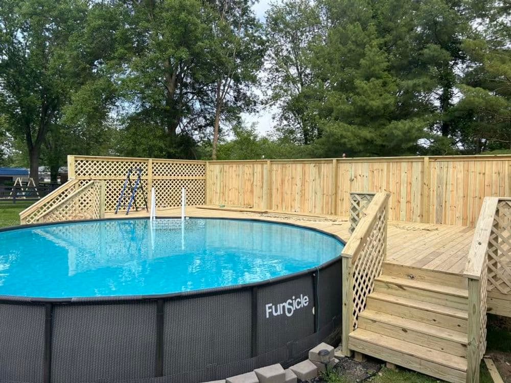 Above-ground swimming pool with wooden deck and railing in a lush green backyard.