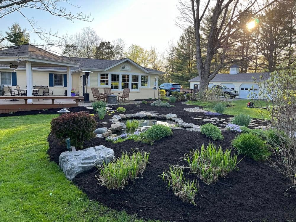 Lush garden with pond, stone features, and patio near a picturesque home at sunset.